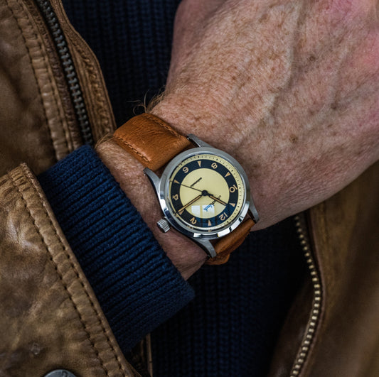 Aeschbach 1957 automatic watch with a tan leather strap worn on wrist with blue sweater and tan leather jacket, emphasizing the sporty yet refined look of the watch.