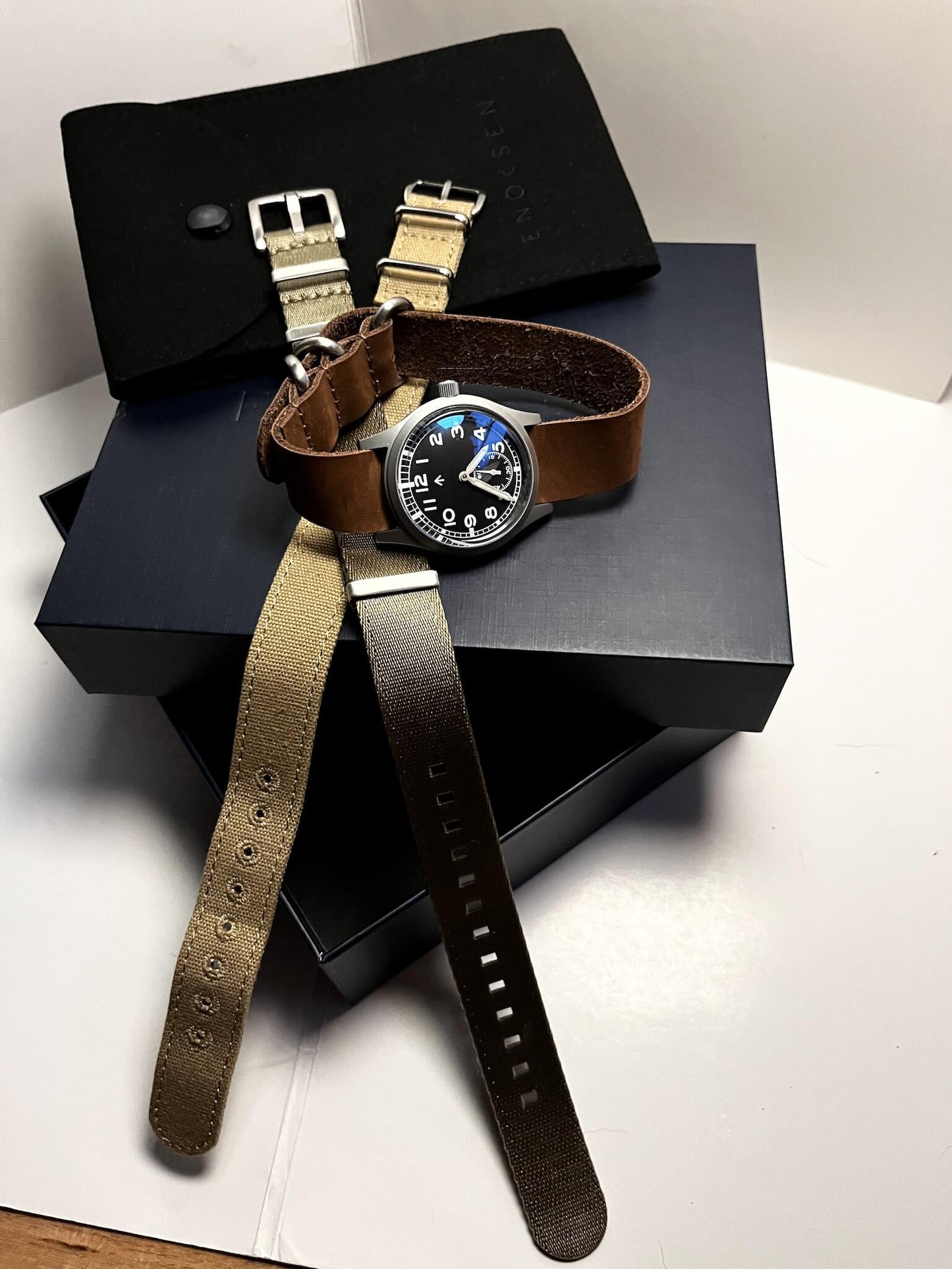 The Enoksen Dirty Dozen swiss edition watch presented on gift box on a premium soft brown leather strap. Also presented is two additional straps that are included with the watch. A Khaki seatbelt NATO strap and a Beige canvas strap giving the customer three looks out of the box.