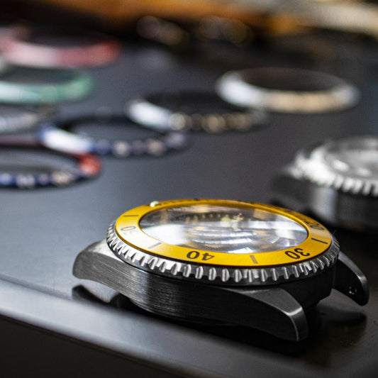 Close-up of a watch with a yellow and black bezel on a dark surface with other watches blurred in the background.