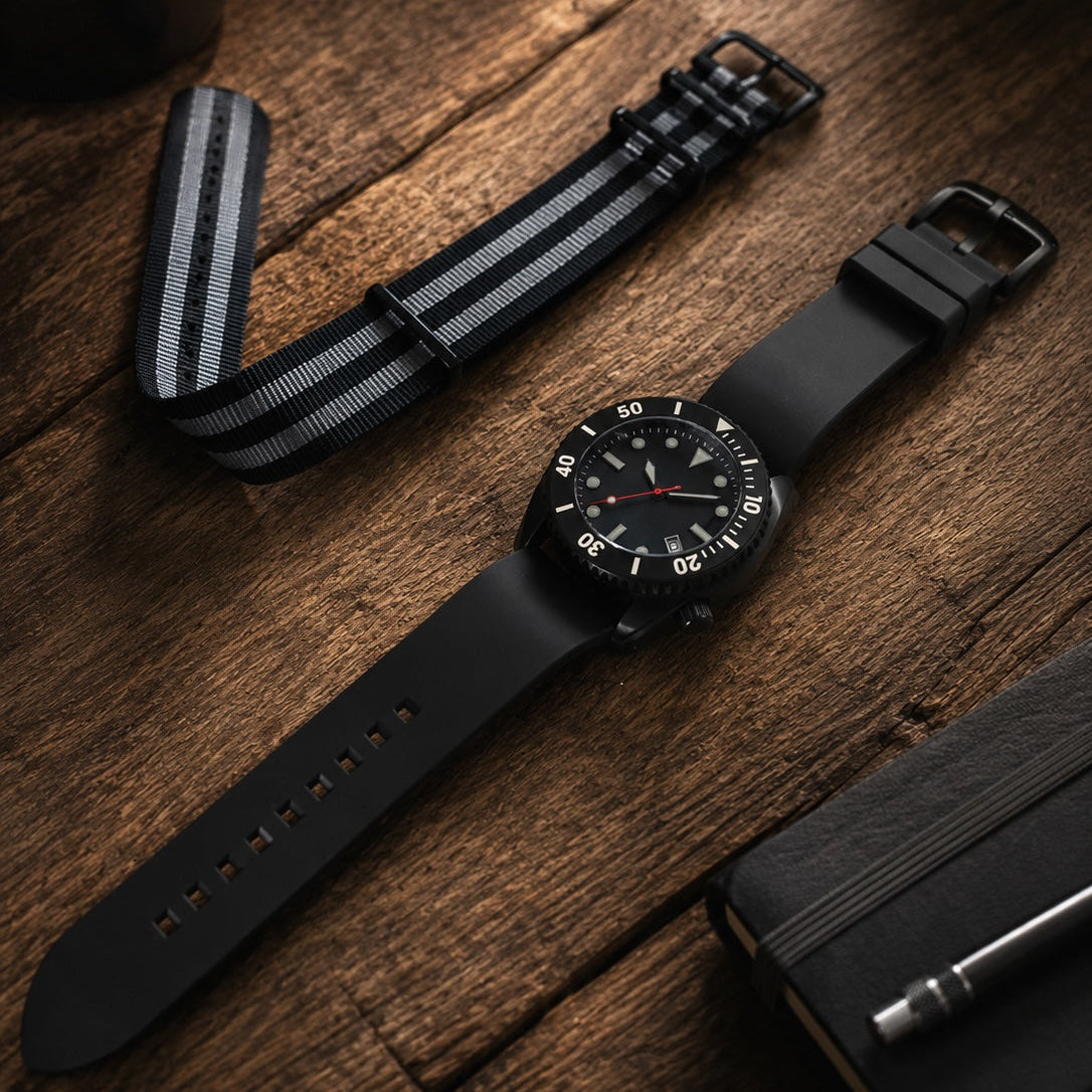 A black diver's watch with a black strap and a striped black-and-white nylon loop band, styled on a rustic wooden tabletop with a coffee cup, notebook, and pen.