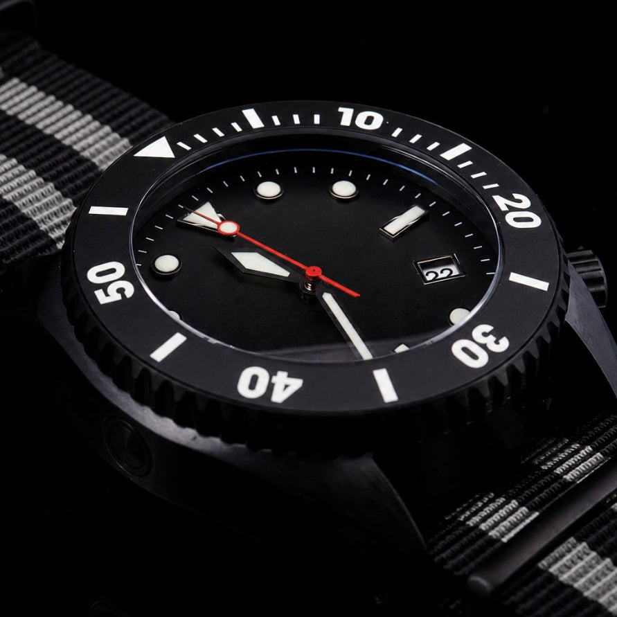 A close-up of a black wristwatch with a black dial, white bezel numerals, and a red second hand, paired with a black and white striped fabric strap, set against a dark background.