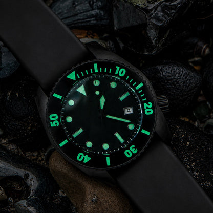 A black wristwatch with a black strap and a glowing green luminescent dial and bezel, photographed resting on dark rocks and shells.