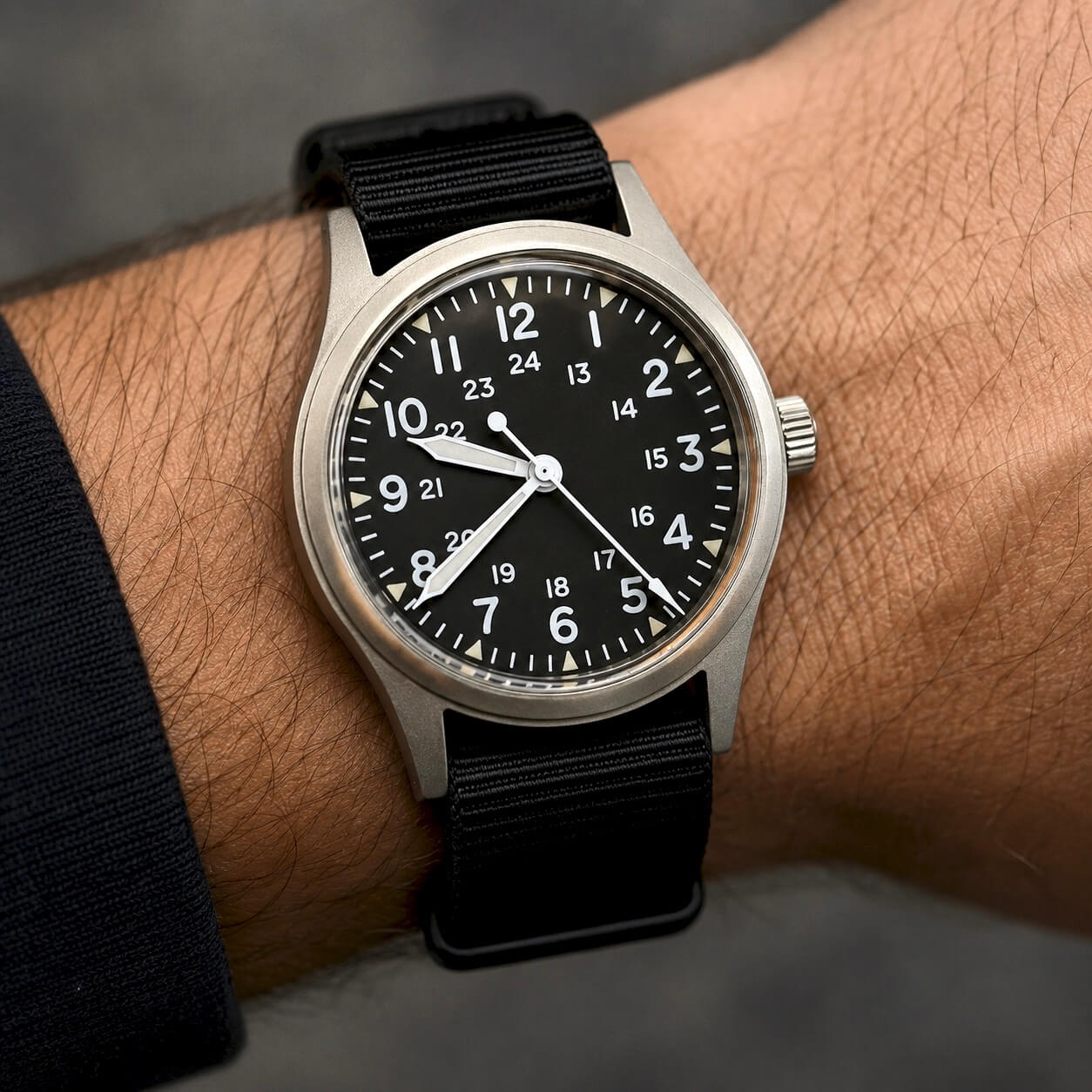 Wrist shot of the Enoksen Fly E03/P mechanical wristwatch with a sandblasted steel case, black dial with white and parchment luminescent markers, 24-hour military dial layout, fixed steel bezel, and black military style nylon strap, on a neutral background.