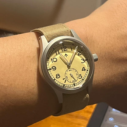 Wrist wearing a watch with a beige canvas strap and cream dial on a wooden surface.