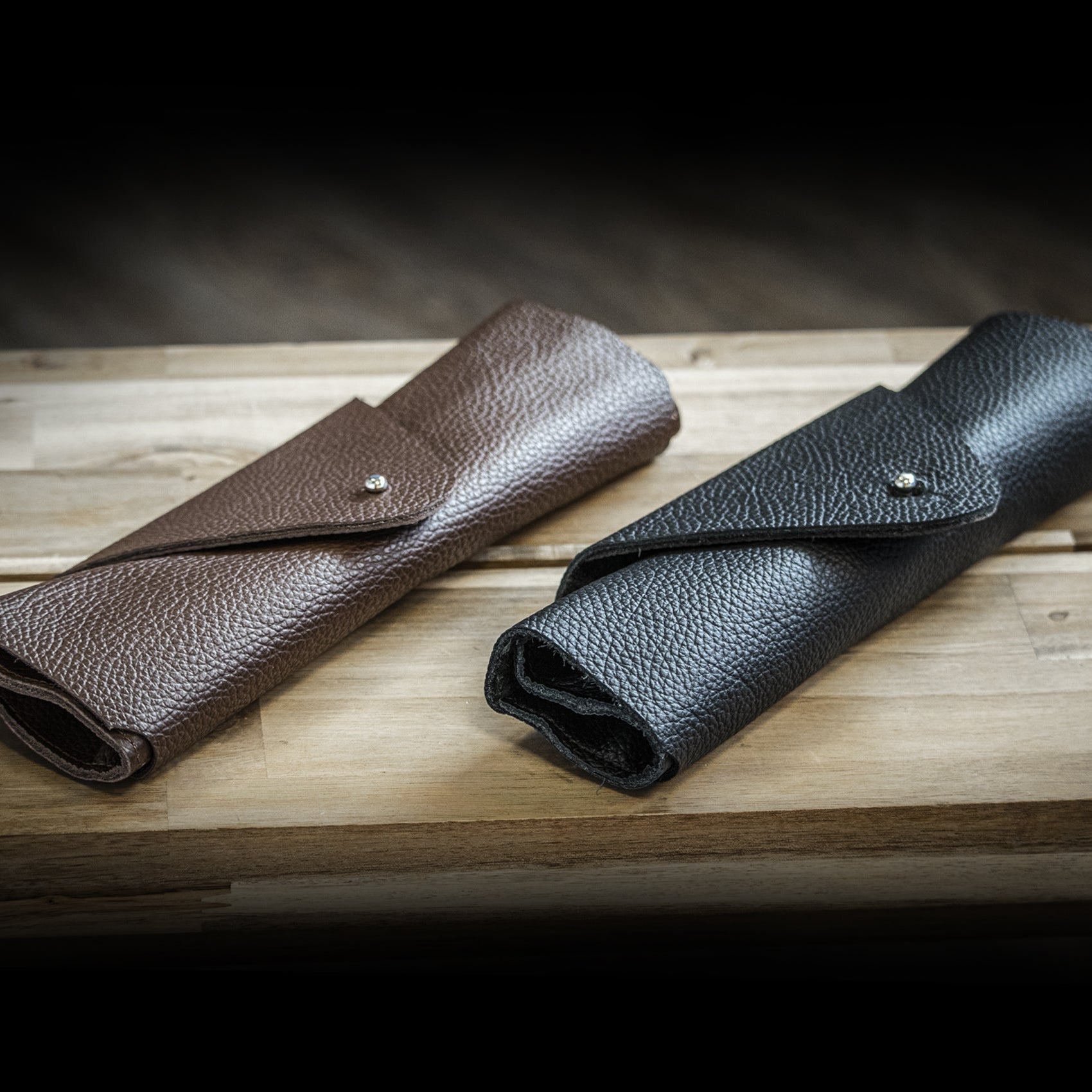Two leather pouches, one brown and one black, on a wooden surface with a dark background.