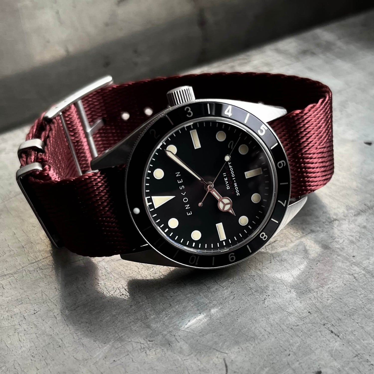Dive II with black dial on burgundy herringbone woven strap, angled wrist-ready view. View on a metal surface with reflection.