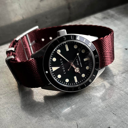 Dive II with black dial on burgundy herringbone woven strap, angled wrist-ready view. View on a metal surface with reflection.