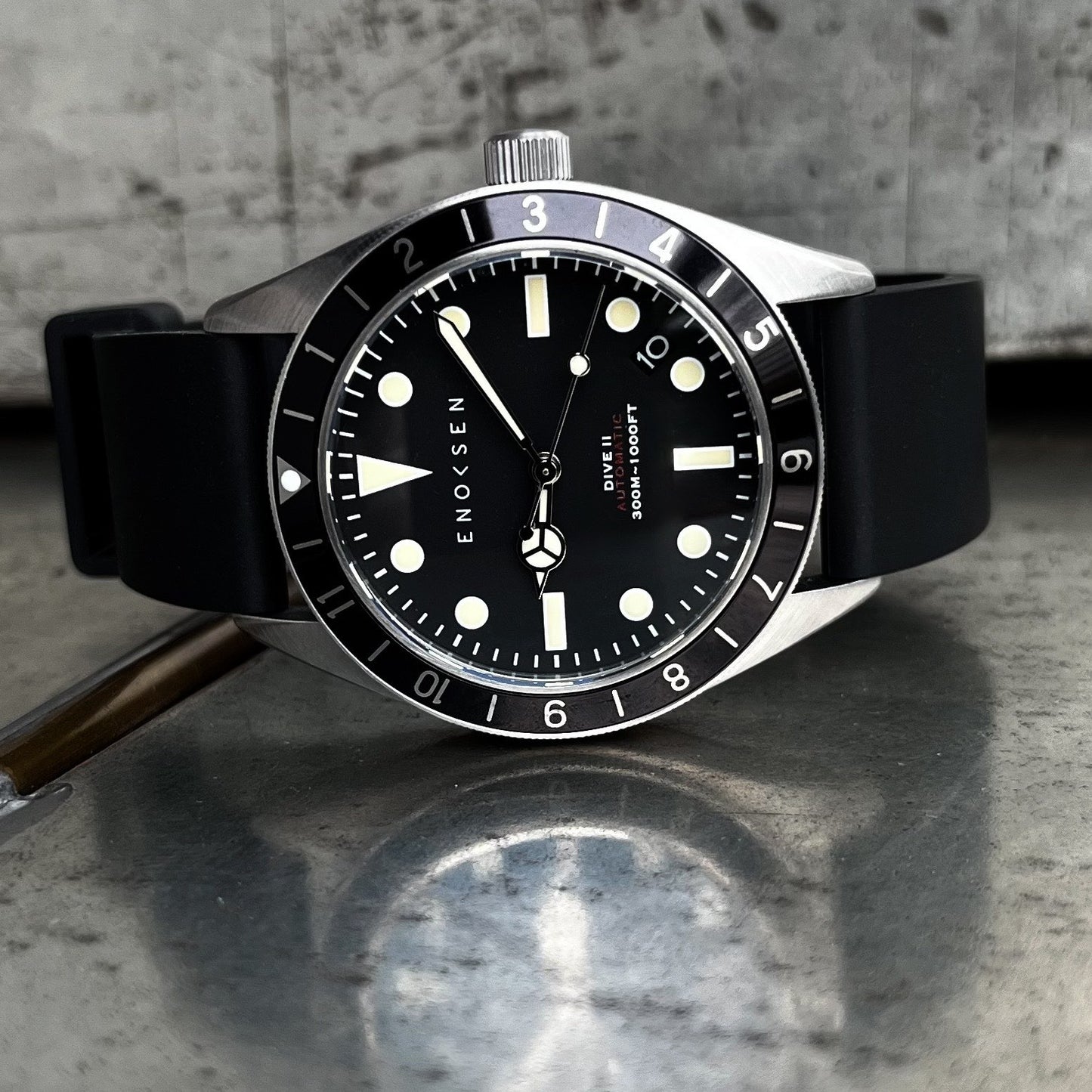 Enoksen Dive II E12/B Skin Diver 39mm with black dial on black rubber strap, front view. View on a metal surface with reflection.