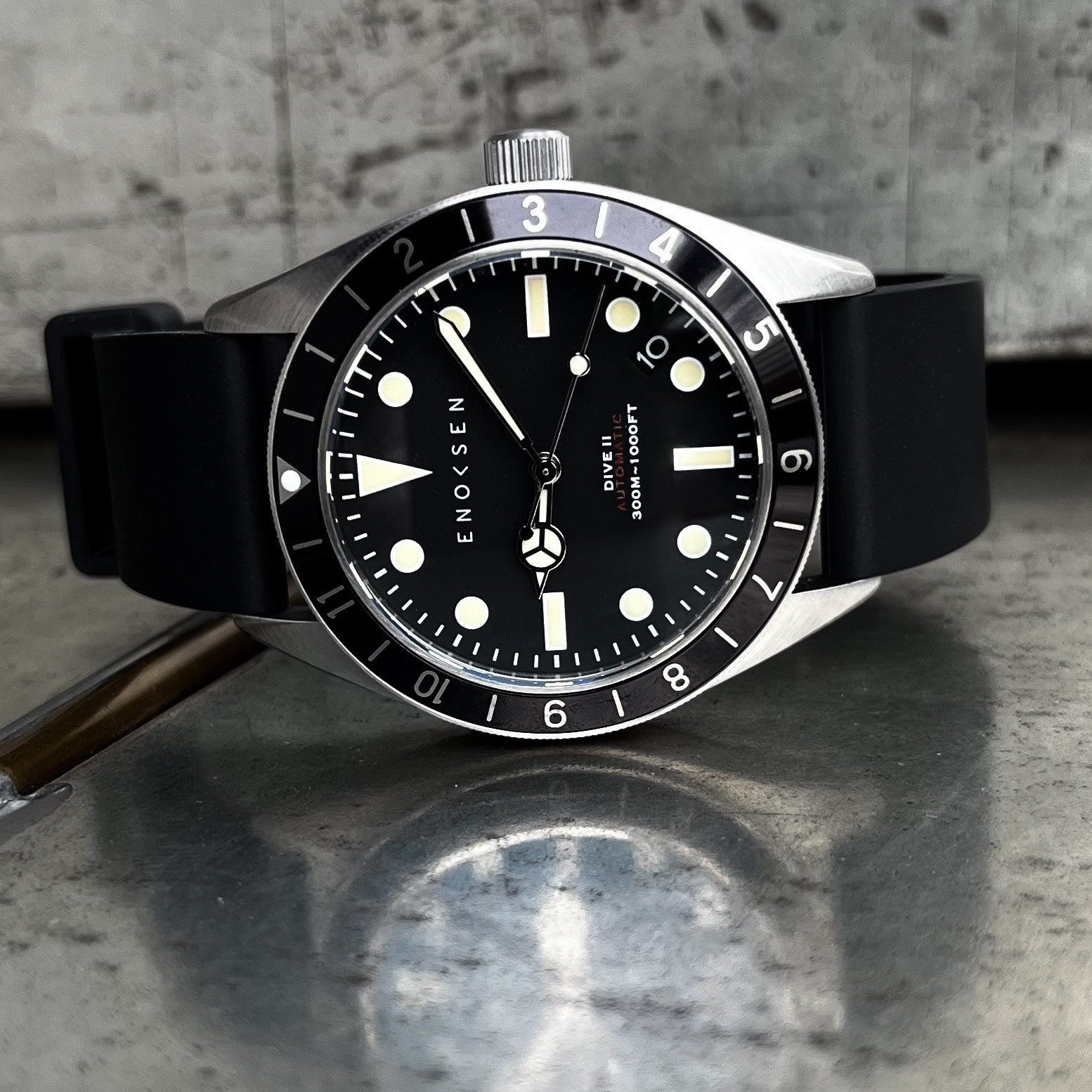 Enoksen Dive II E12/B Skin Diver 39mm with black dial on black rubber strap, front view. View on a metal surface with reflection.