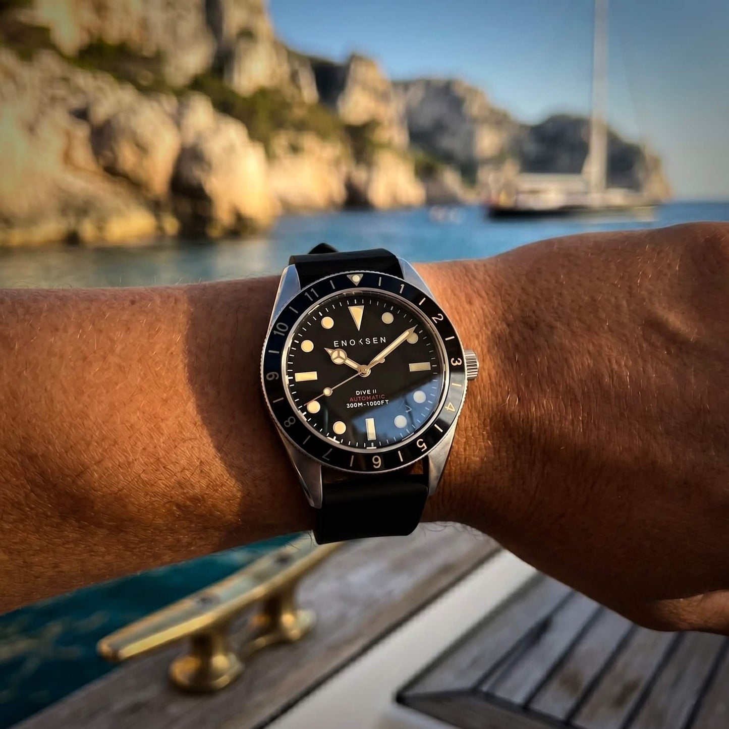 Enoksen Dive II E12/B black dial worn on a wrist size 19cm, showcasing the proportions of the watch and the classic skin-diver profile. Background is on a boat in rocky sunny landscape.