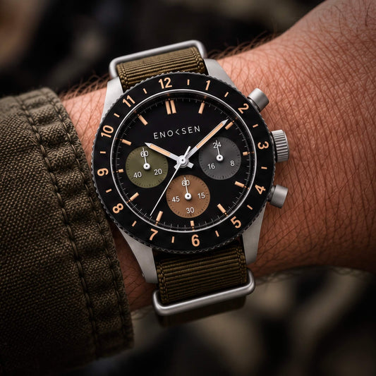 A close-up of a wrist wearing an Enoksen FIELD 002 chronograph watch with a black dial, silver-toned case, and olive green fabric strap.