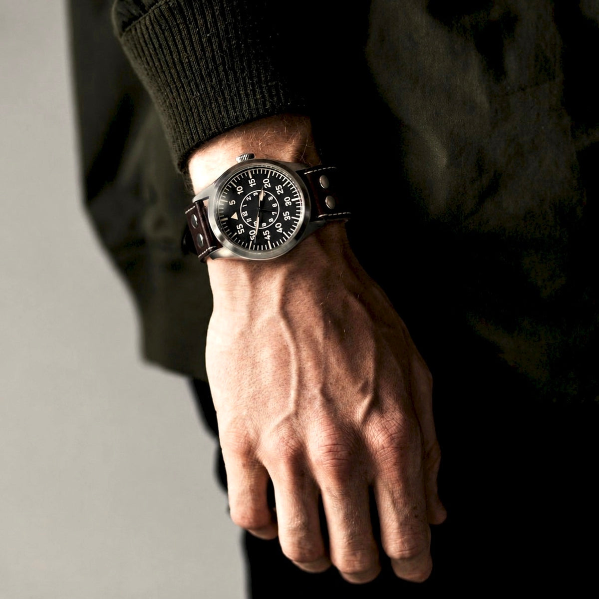 Enoksen FLY E03/B pilot watch on a man's wrist with a dark sleeve, showing the black dial and brown leather strap.