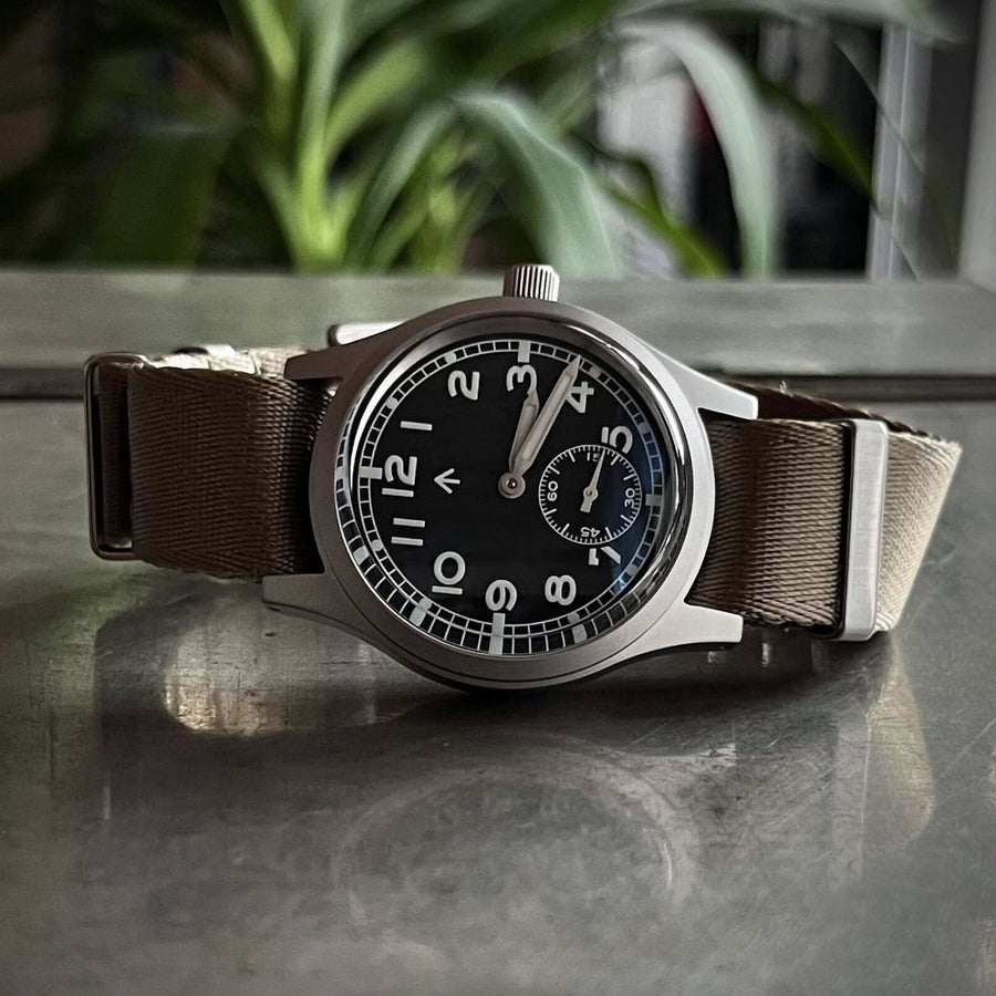 Front view of the Enoksen Fly E03/S automatic 36mm Dirty Dozen field watch with a black dial on a khaki NATO strap. Shown on a metal surface with reflection.