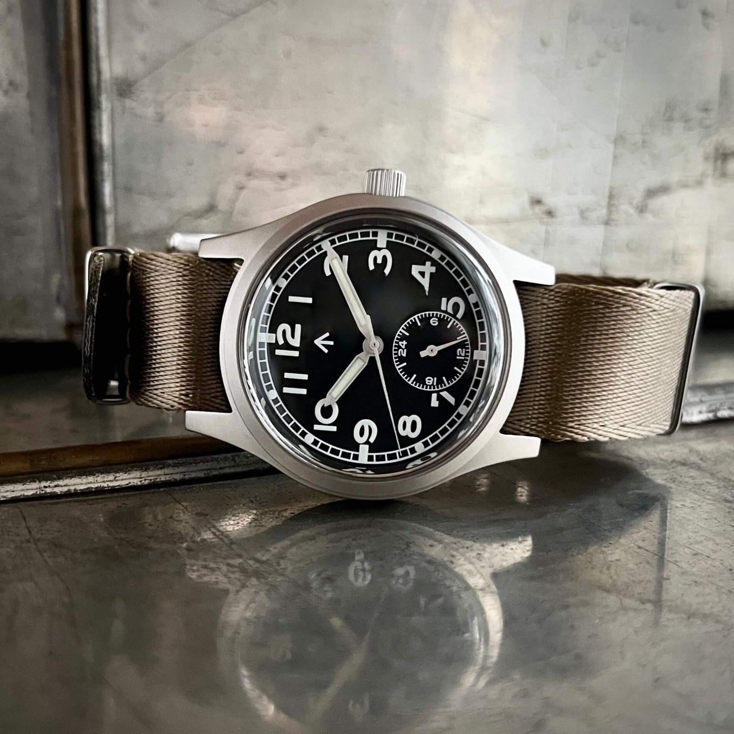 Enoksen fly E03/H black dial Dirty Dozen watch on a Khaki seatbelt NATO strap on metal box with reflection.