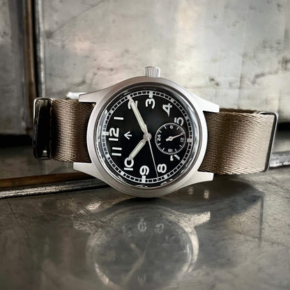 Enoksen fly E03/H black dial Dirty Dozen watch on a Khaki seatbelt NATO strap on metal box with reflection.