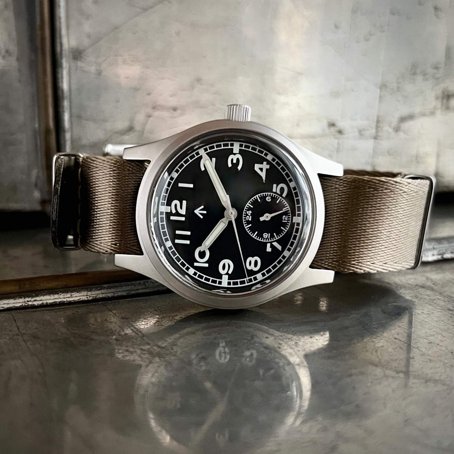 Front view of the Enoksen Fly E03/H automatic 36mm Dirty Dozen field watch with a black dial on a khaki NATO strap. Shown on a metal surface with reflection.