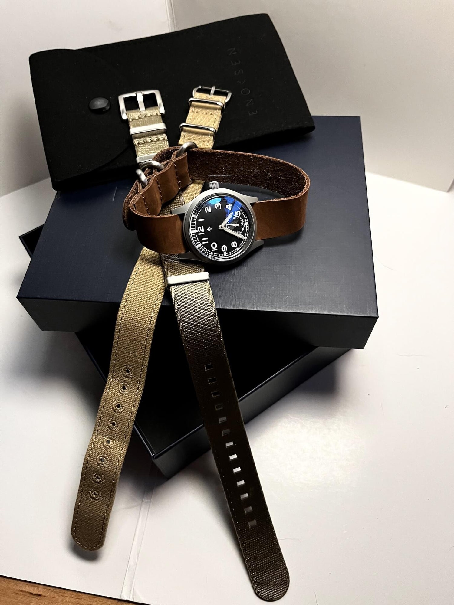 The Enoksen Dirty Dozen swiss edition watch presented on gift box on a premium soft brown leather strap. Also presented is two additional straps that are included with the watch. A Khaki seatbelt NATO strap and a Beige canvas strap giving the customer three looks out of the box.
