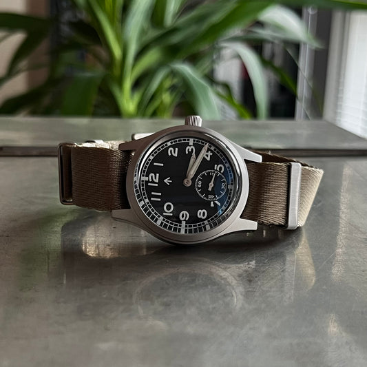 Fly E03/S with khaki NATO double pass strap. Angled wrist-ready view on a metal surface with reflection. Green plant in background