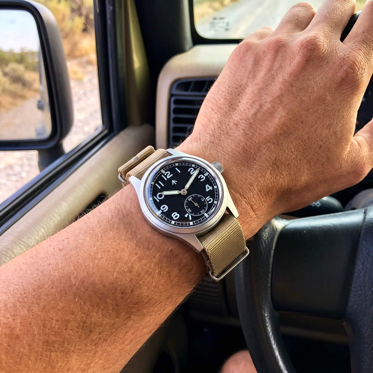 Enoksen Fly E03/S watch with a black dial and khaki NATO strap worn on a wrist by a man sitting in a Jeep.