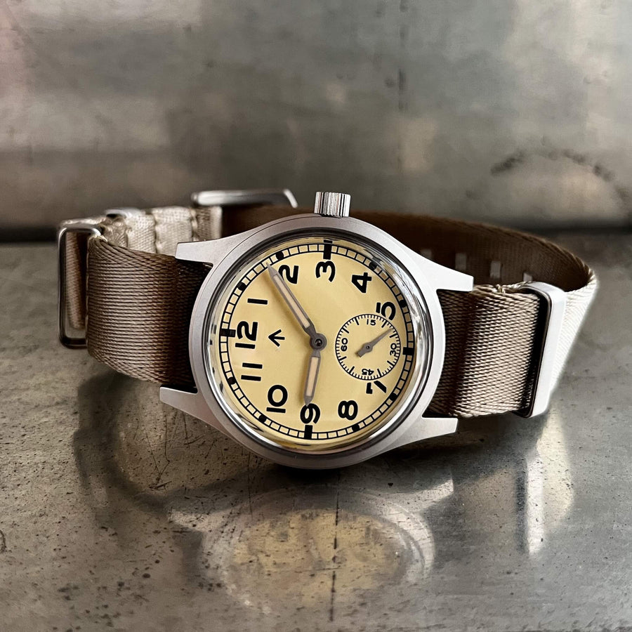 Front view of the Enoksen Fly E03/T automatic 36mm Dirty Dozen field watch with a cream dial on a khaki NATO strap. Shown on a metal surface with reflection.