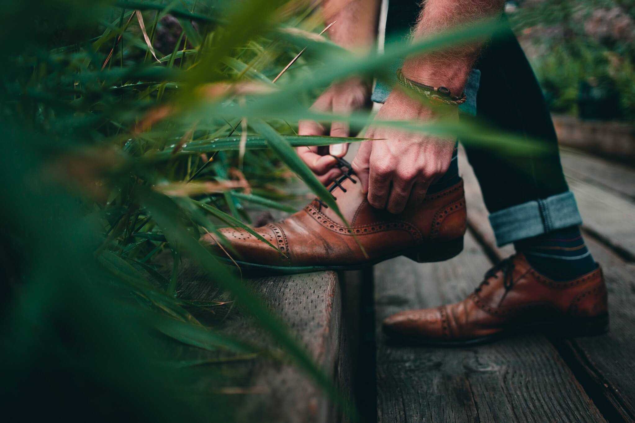 Person adjusting brown leather shoes on a wooden bench with greenery in the foreground