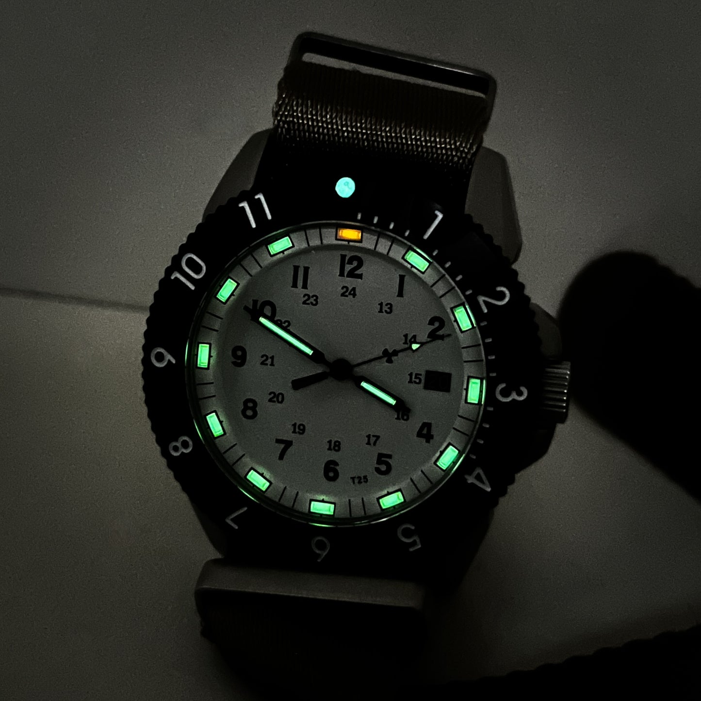 Enoksen Tactic E09/AQ Swiss Quartz wristwatch with a sandblasted steel case, white dial with green and orange Tritium luminescent markers, black rotating 12-hour bezel, and khaki military style nylon strap, on a stone surface.
