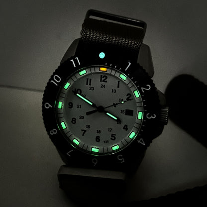 Enoksen Tactic E09/AQ Swiss Quartz wristwatch with a sandblasted steel case, white dial with green and orange Tritium luminescent markers, black rotating 12-hour bezel, and khaki military style nylon strap, on a stone surface.