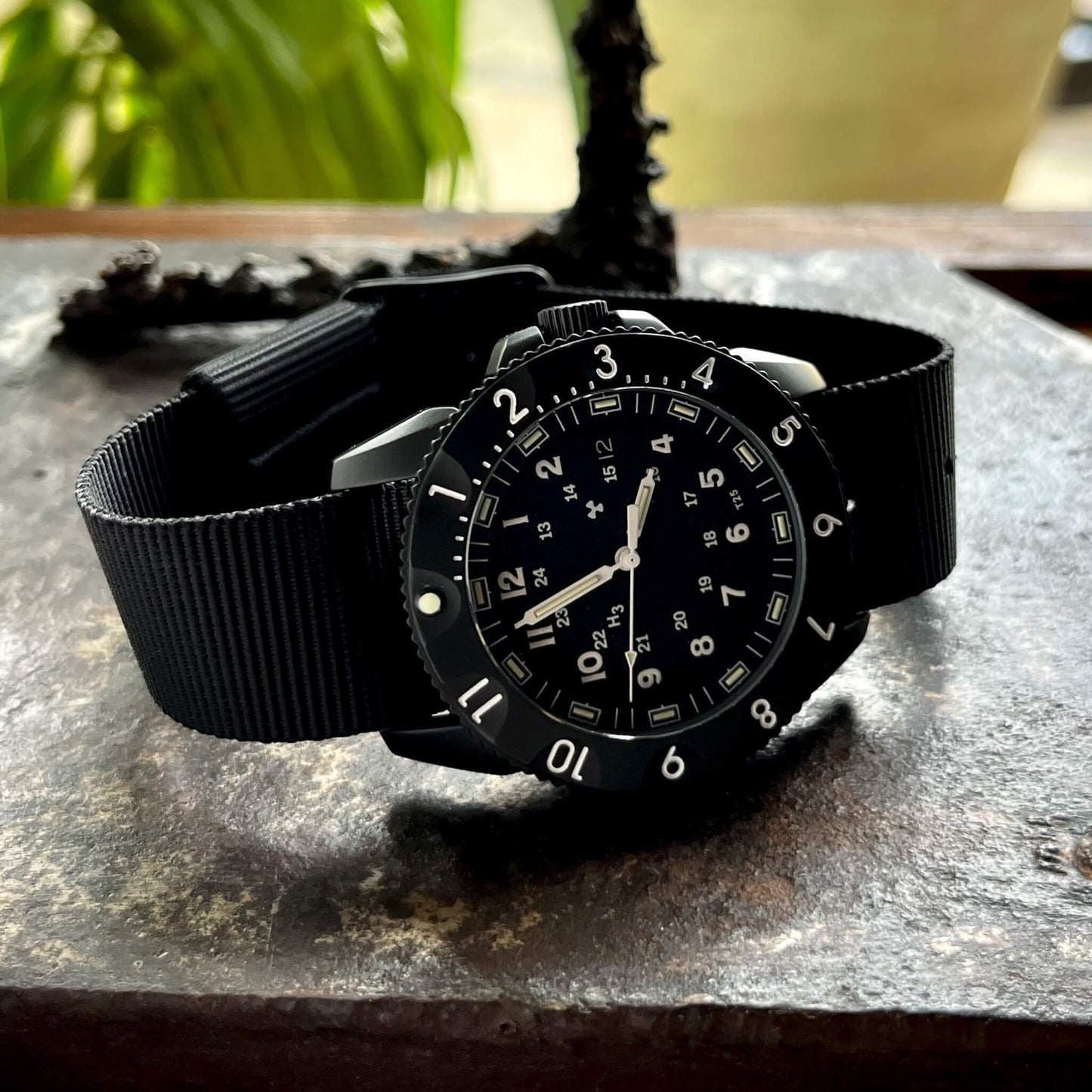 Enoksen Tactic E09/BQ Swiss Quartz wristwatch with a black dial, black rotating 12-hour bezel, and black military style nylon strap, on a stone surface.