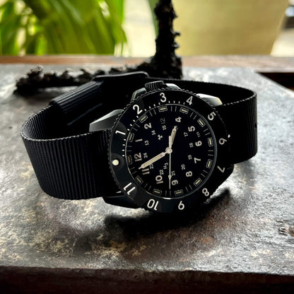 Enoksen Tactic E09/BQ Swiss Quartz wristwatch with a black dial, black rotating 12-hour bezel, and black military style nylon strap, on a stone surface.