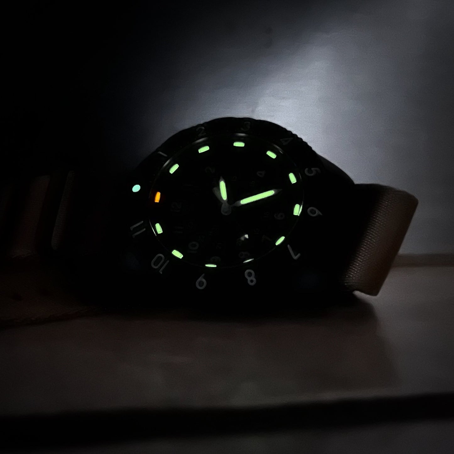 Enoksen Tactic E09/BQ Swiss Quartz wristwatch with a sandblasted steel case, white dial with green and orange Tritium luminescent markers, black rotating 12-hour bezel, and khaki military style nylon strap, in nearly complete darkness, on a metal surface.