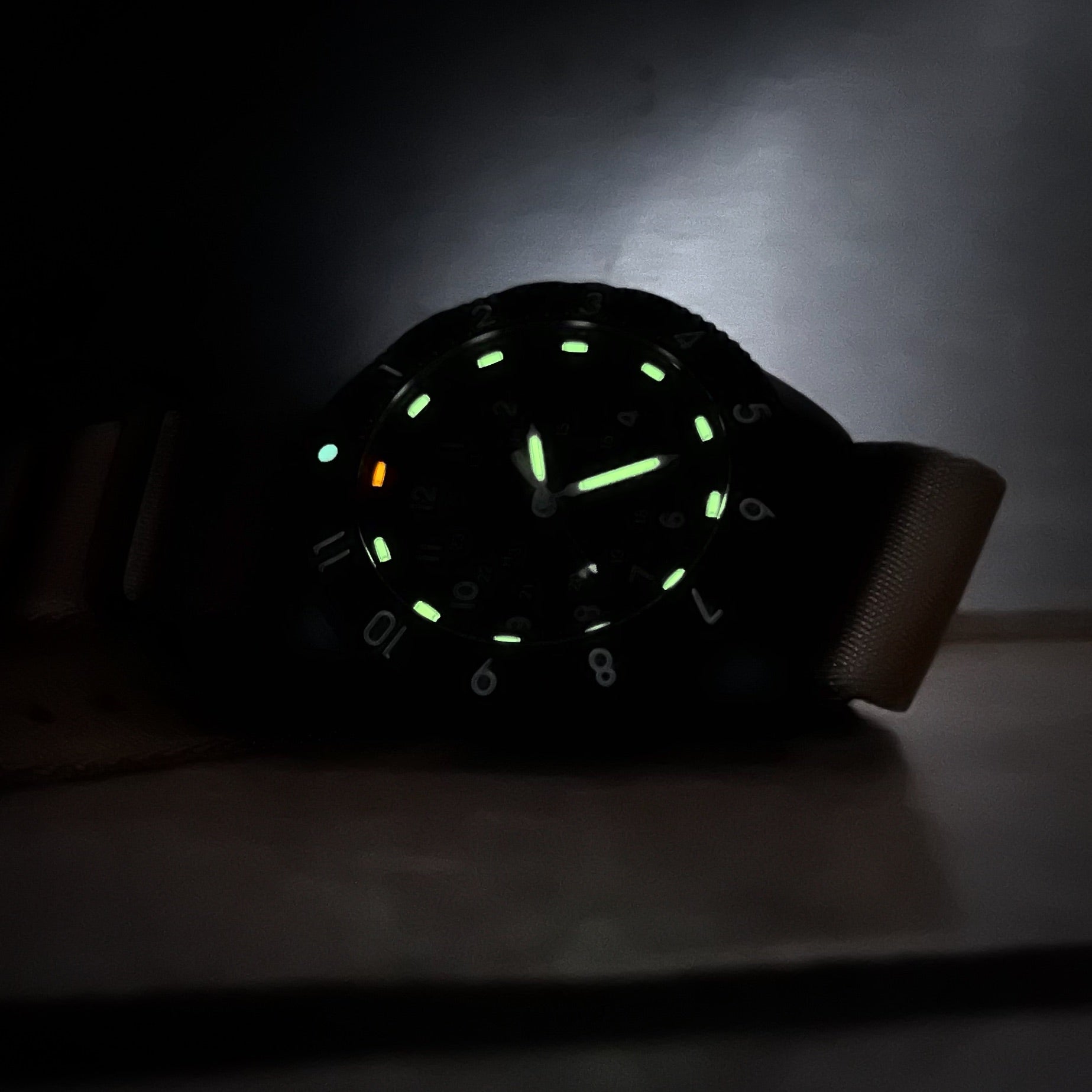 Enoksen Tactic E09/BQ Swiss Quartz wristwatch with a sandblasted steel case, white dial with green and orange Tritium luminescent markers, black rotating 12-hour bezel, and khaki military style nylon strap, in nearly complete darkness, on a metal surface.
