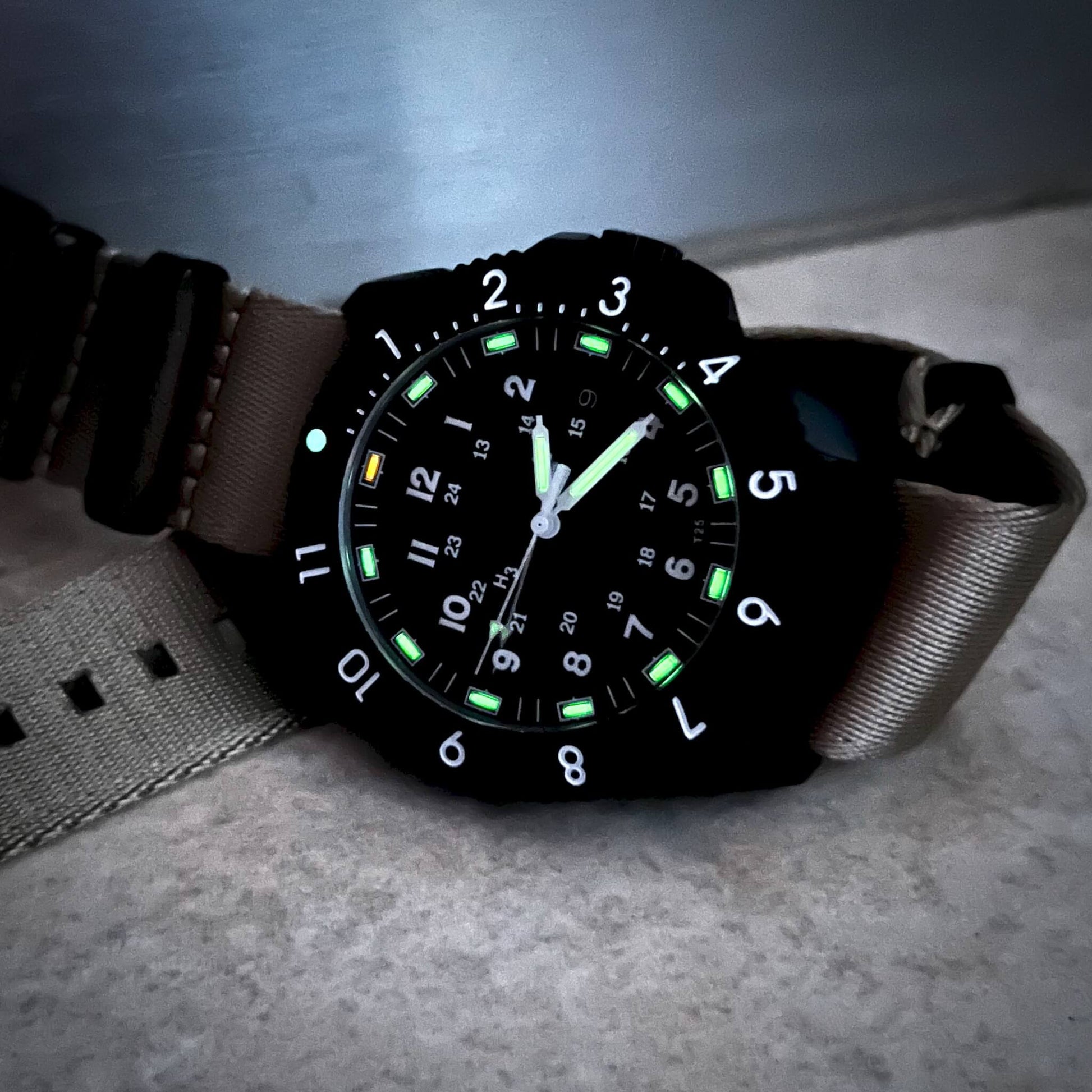 Enoksen Tactic E09/BQ Swiss Quartz wristwatch with a sandblasted steel case, black dial with green and orange Tritium luminescent markers, black rotating 12-hour bezel, and khaki military style nylon strap, in dimmed light, on a metal surface.