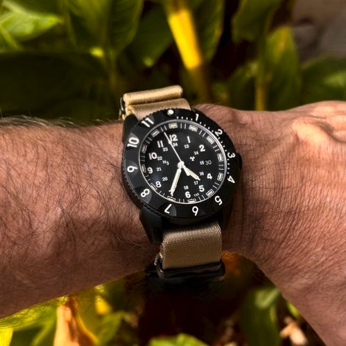 Enoksen Tactic E09/BQ Swiss Quartz wristwatch with a black dial, black rotating 12-hour bezel, and Khaki seatbelt nylon NATO strap with blacked out metal dressers, on a wrist, blurred natural background.