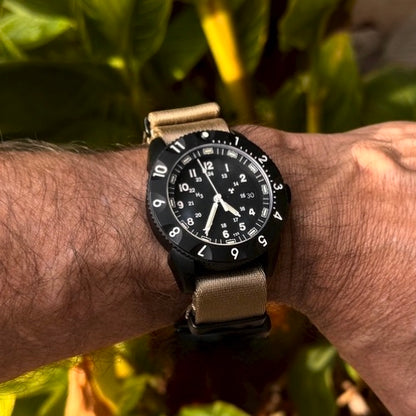Enoksen Tactic E09/BQ Swiss Quartz wristwatch with a black dial, black rotating 12-hour bezel, and Khaki seatbelt nylon NATO strap with blacked out metal dressers, on a wrist, blurred natural background.