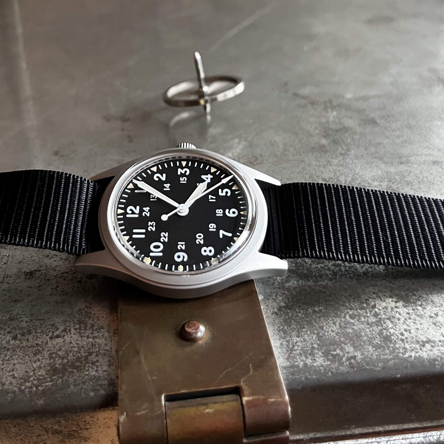 Enoksen Fly E03/P mechanical wristwatch with a sandblasted steel case, black dial with white and parchment luminescent markers, 24-hour military dial layout, fixed steel bezel, and black military style nylon strap, on a metal surface.
