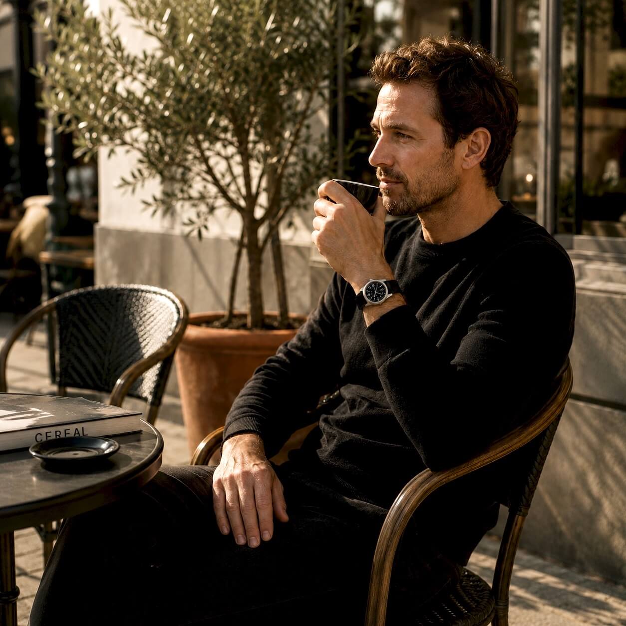 Enoksen Fly E03/P mechanical wristwatch with a sandblasted steel case, black dial with white and parchment luminescent markers, 24-hour military dial layout, fixed steel bezel, and black military style nylon strap, worn by a man drinking coffee in front of a cafe with an olive tree in the background.